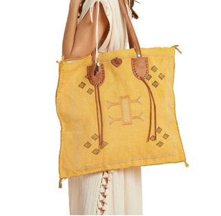 A yellow woven tote bag with brown leather handles and embroidered geometric details, shown being carried by a model wearing a cream dress against a white background.