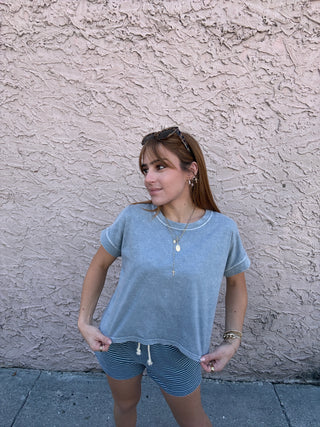 Woman wearing a blue top and striped shorts standing against a textured wall.