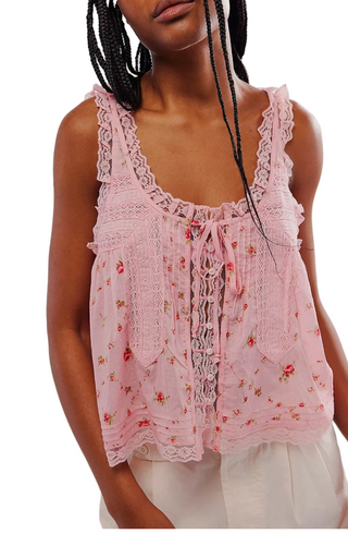 A close-up of a model wearing a light pink sleeveless top with delicate floral embroidery, lace trim, and a front tie closure, paired with white bottoms against a plain light background.