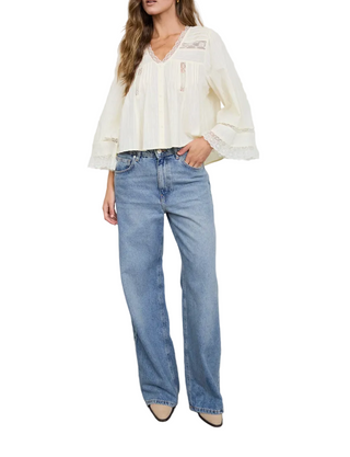 A woman modeling a cream-colored blouse with ruffled sleeves and a lace-trimmed V-neck, paired with light-wash blue wide-leg jeans and beige pointed-toe heels, standing against a white wall.