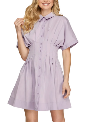 A woman wearing a light lavender short-sleeve button-up shirt dress with a smocked waist and flared skirt, shown against a plain light gray background.
