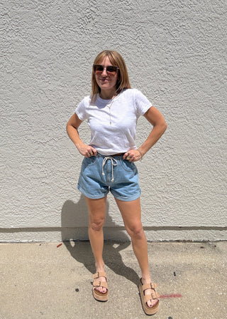 Person wearing a white shirt and denim shorts standing on a sidewalk.