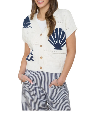 A woman wearing a cream short-sleeve knit cardigan with navy blue seashell and wave motifs, paired with blue and white striped pants.