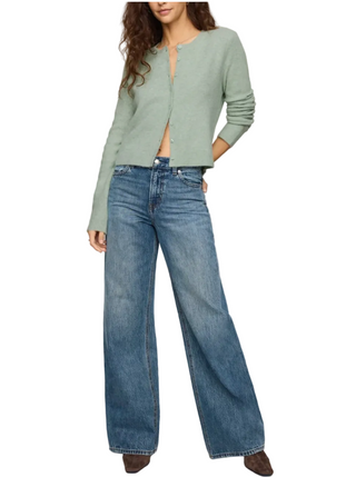 A model wearing a sage green cropped cardigan and wide-leg blue jeans, standing against a neutral light beige studio background.