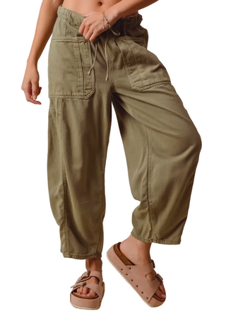 A person wearing loose olive green cargo-style pants with a drawstring waist and large front cargo pockets, paired with beige platform sandals against a plain beige studio background.