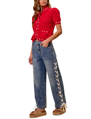 A model wearing a vibrant red short-sleeve knit top with white contrast stitching and high-waisted, wide-leg blue jeans featuring white lace-up detailing down the outer side, paired with brown platform sandals, set against a solid beige background.