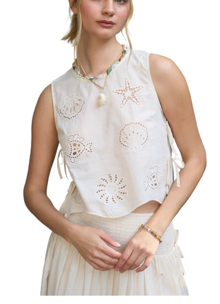 Woman wearing a white sleeveless top with decorative cut-out patterns against a gray wall.