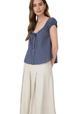 A woman modeling a navy and white gingham check top with cap sleeves and a ruffled front, paired with a cream pleated skirt, against a light gray studio background.