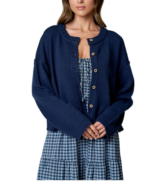 A woman wearing a navy blue knit cardigan with tan buttons over a blue and white plaid dress, shown from the nose down against a light gray studio background.
