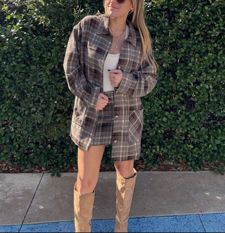 A woman wearing an oversized brown and beige plaid flannel shirt dress over a white tank top, paired with tan knee-high boots, standing outdoors in front of a green hedge.