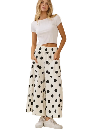 A woman modeling a white ribbed short-sleeve crop top and wide-leg pants with a black polka dot pattern, paired with white sneakers, against a plain beige studio background.
