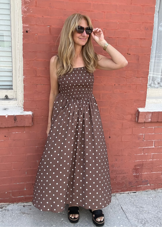 woman wearing a brown dress with white polka dots against a brick wall background