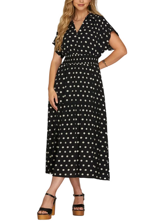 A woman models a black midi dress with a white polka dot pattern, featuring flutter sleeves, a V-neckline, and a matching fabric belt at the waist. She is wearing black platform sandals against a neutral gray studio background.