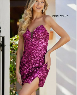 A woman models a bright magenta grape sequined strapless mini dress with a sweetheart neckline and a small front slit. The brand name 'PRIMAVERA' is printed in black text on the upper right side of the image.