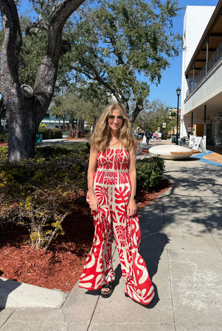 Woman in a red and white patterned outfit standing on a sidewalk with trees and buildings in the background.