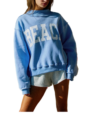 A model wearing a light blue crewneck sweatshirt with 'BEACH' printed in white varsity letters across the back, paired with light gray shorts, set against a rocky outdoor background.