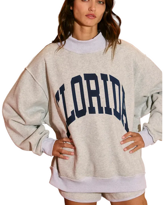 A woman wearing an oversized light heather gray sweatshirt with 'FLORIDA' printed in navy blue arched letters across the chest, paired with matching light lavender shorts, standing in a room with orange walls and floral wallpaper.