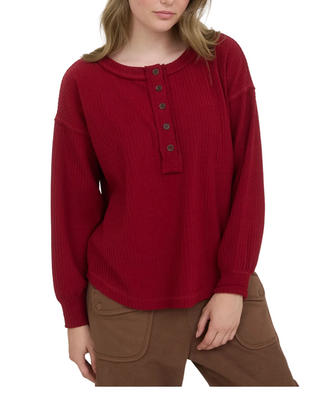 A woman wearing a ribbed long-sleeve henley top in deep burgundy red with dark buttons, paired with brown pants, against a plain light background.