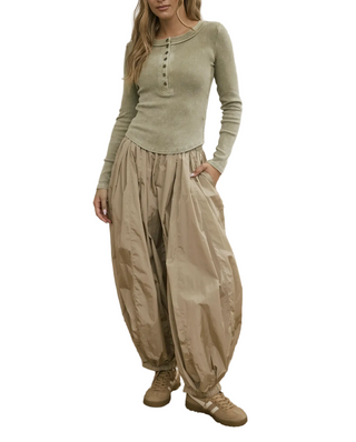 A woman stands in a cozy room wearing a light sage green long-sleeve henley top and loose beige harem-style pants with side pockets, paired with beige sneakers. A floral sofa, small TV, and woven rug are visible in the background.