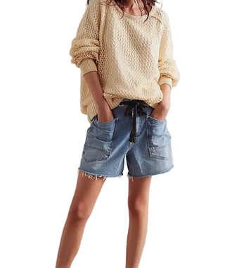 A person wearing a cream-colored chunky knit sweater and light-wash blue denim shorts with front patch pockets and a frayed hem, standing against a plain off-white wall.