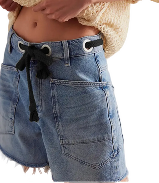 Close-up of a person wearing light blue denim shorts with a black drawstring waist and white metal grommets, paired with a cream crochet top against a white background.