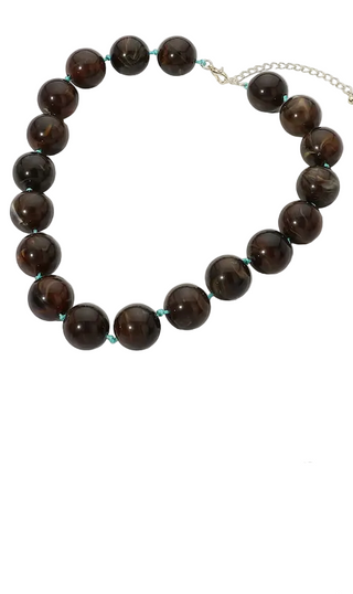 Two beaded necklaces displayed on a white marble surface: one with dark brown polished beads and one with light blue matte beads, each with a small gold chain extender. A partial hand is visible at the right edge in both panels.