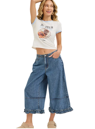 A woman modeling a white ringer crop top with a graphic print and high-waisted, wide-leg blue denim culottes with ruffled hems, paired with beige slide sandals, against a plain light background.