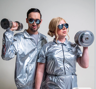 Two people in silver reflective outfits lifting weights against a plain background