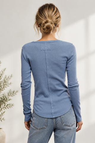 A woman viewed from the back wearing a fitted long-sleeve top in dusty light blue, paired with olive green pants, against a plain light gray studio background.