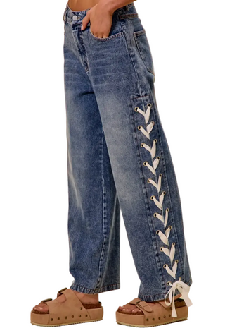 A person wearing wide-leg blue denim jeans with decorative white lace-up detailing running down the outer side of each leg, paired with brown platform sandals against a plain beige studio background.