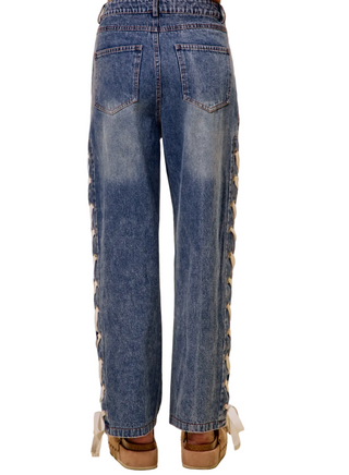 Rear view of a person wearing blue denim jeans with lace-up side detailing and white laces at the ankles, paired with tan wedge sandals, against a beige studio background.