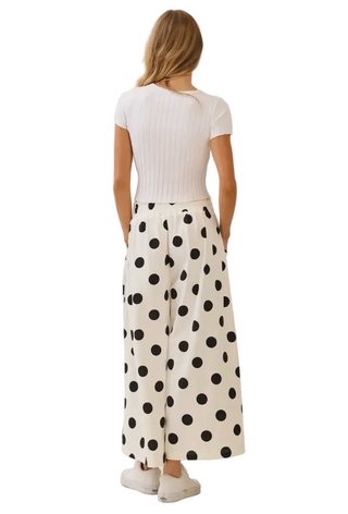 A woman is shown from behind wearing a white ribbed short-sleeve top and white wide-leg pants with large black polka dots, paired with white sneakers, against a plain cream background.