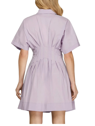 A woman shown from the back wearing a short-sleeve lavender shirt dress with a pleated back and flared skirt, photographed against a plain light gray studio background.