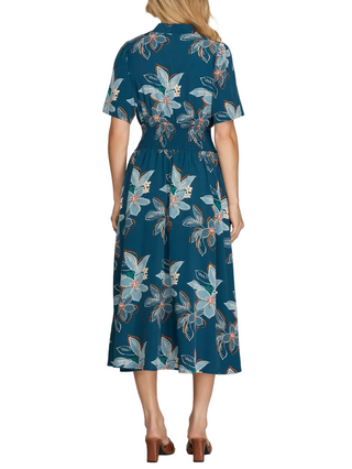 A woman modeling a short-sleeve midi dress from the back, featuring a teal blue base with a light blue and white floral pattern and small orange accents. She is wearing brown block-heel mules against a neutral gray studio background.