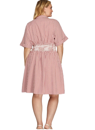 A woman modeling a red and white gingham short-sleeve dress from the back, featuring a gathered waist and a matching gingham belt, set against a plain light gray studio background.