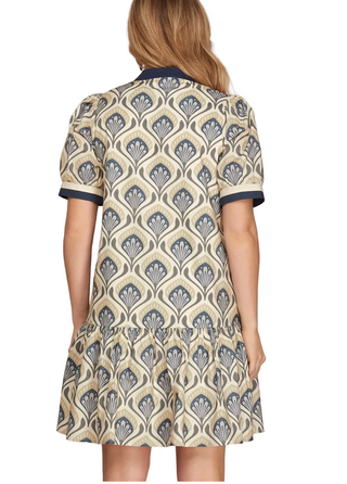 A woman shown from the back wearing a short-sleeve dress with a beige base and a repeating navy blue and gray geometric fan pattern, set against a solid light gray background.
