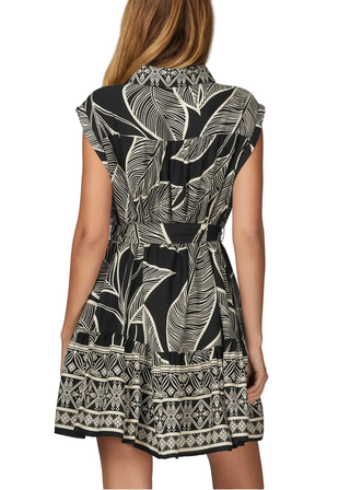 A woman shown from the back wearing a sleeveless collared dress with a black and off-white tropical leaf print and a geometric patterned hem, set against a plain light gray studio background.
