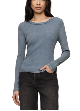 A woman wearing a fitted, long-sleeve ribbed knit top in a muted dusty blue color, paired with dark gray jeans, against a solid light beige background.