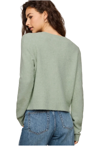 A woman shown from the back wearing a light sage green knit sweater and blue jeans against a plain light beige studio background.