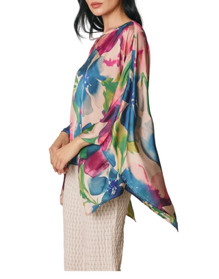 A woman wearing a lightweight floral print top with wide draped sleeves and a draped hem, paired with a textured cream skirt, shown against a neutral studio background.