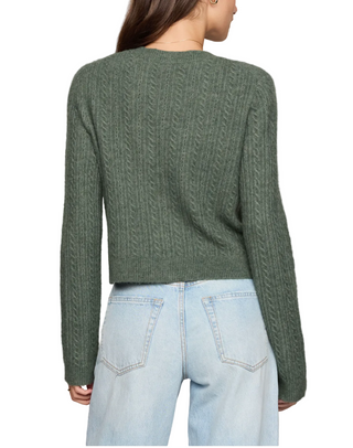 A woman shown from the back wearing a cropped, long-sleeve cable-knit sweater in sage green, paired with light-wash blue jeans, against a solid light beige background.