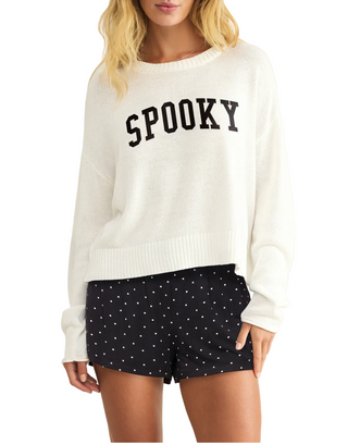 A woman wearing a cream knit sweater with 'SPOOKY' printed in black across the chest, paired with black shorts covered in small white polka dots, against a light beige studio background.