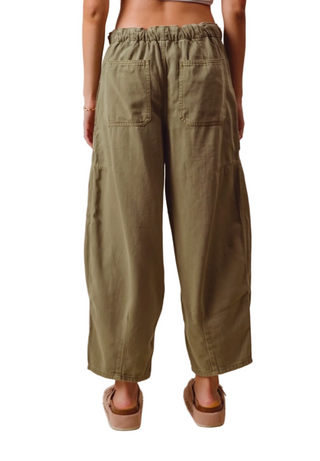 A person shown from the back wearing loose, wide-leg olive green pants with an elastic waistband and two large back patch pockets, paired with tan sandals against a plain beige studio background.