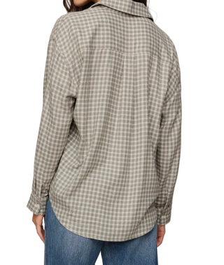 A person shown from the back wearing an oversized long-sleeve button-up shirt with a muted green and beige gingham check pattern, paired with blue jeans, against a plain light beige background.