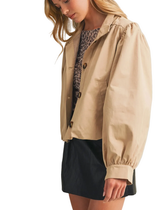 A woman wearing a beige cropped utility jacket with balloon sleeves over a leopard-print top and a black skirt, photographed against a plain light beige studio background.