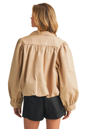 A woman shown from the back wearing a beige long-sleeve blouse with gathered detailing and black shorts, standing against a plain light beige studio background.