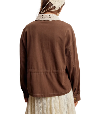 A person shown from the back wearing a brown long-sleeve jacket with a cream lace collar and a matching cream lace skirt, against a plain light beige studio background.