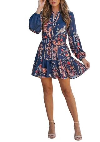 A woman models a navy blue long-sleeve mini dress with a colorful floral and bird print, standing against a textured beige concrete wall.
