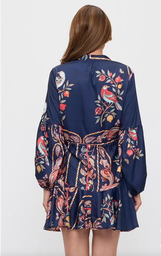 A woman viewed from the back wearing a navy blue long-sleeve mini dress with a colorful bird and floral print, photographed against a light gray studio background.