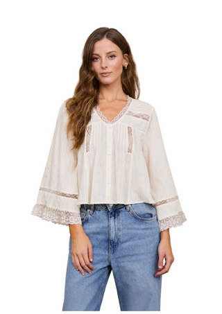 A woman wearing a cream-colored, long-sleeve bohemian-style blouse with lace trim and blue jeans, standing against a rustic textured stucco wall.